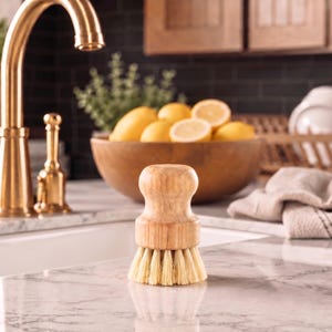 Puede incluir: Un cepillo de madera para lavar platos con cerdas de color claro est&aacute; sobre una encimera de m&aacute;rmol. Un grifo dorado y un cuenco de madera lleno de limones est&aacute;n en el fondo. Tambi&eacute;n se ve una toalla de lino y un escurreplatos.