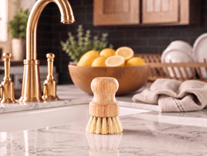 Puede incluir: Un cepillo de madera para lavar platos con cerdas de color claro est&aacute; sobre una encimera de m&aacute;rmol. Un grifo dorado y un cuenco de madera lleno de limones est&aacute;n en el fondo. Tambi&eacute;n se ve una toalla de lino y un escurreplatos.