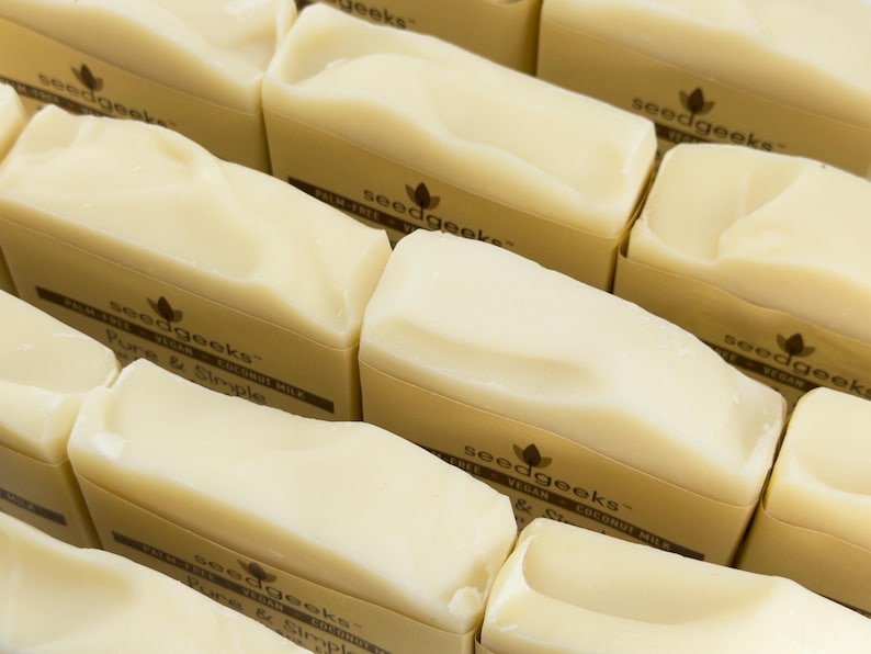 May include: Multiple rectangular bars of off-white soap arranged in a grid. Each bar is wrapped in a tan paper band featuring the text seedgeeks, palm-free, vegan, coconut milk, and Pure & Simple printed in a dark brown font.