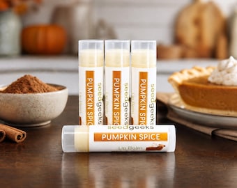 May include: Four cylindrical lip balm tubes with clear caps and white labels. Three tubes stand vertically, and one lies horizontally in front. The labels feature the text seedgeeks PUMPKIN SPICE Lip Balm with an orange and white color scheme.