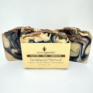 May include: Three bars of handcrafted soap with a swirling pattern of black, brown, and cream colors. The soap is labeled "Sandalwood Patchouli" and is palm-free and vegan. Each bar weighs 4 oz - 5 oz.