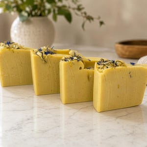 May include: Four rectangular bars of pale yellow soap arranged in a row. Each bar features a smooth, solid texture with visible flecks and is topped with a decorative scattering of dried yellow and blue flower petals across the top edge.