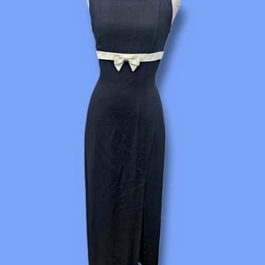 May include: A sleeveless, black dress with a white bow detail at the waist. The dress has a slit up the front and is displayed on a mannequin. The bow is centered on a white band that wraps around the waist.