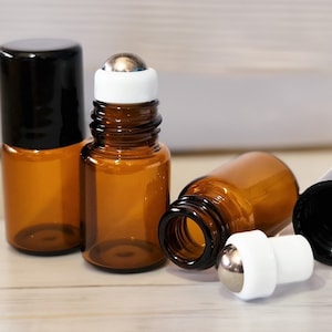 May include: A set of amber glass roller bottles with black screw-on caps and white plastic inserts featuring a metal ball applicator. Two bottles are standing upright, one is lying on its side, and one separate roller insert is placed nearby.