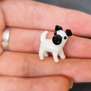 May include: A miniature glass figurine of a dog with a white body, black ears, and a black snout. The small animal sculpture features a smooth, glossy finish and is depicted in a standing position with its tail pointed upward and alert.