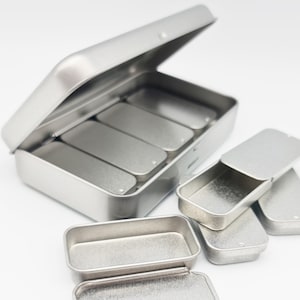 Peut inclure: Ensemble de petites boîtes métalliques rectangulaires argentées. Une grande boîte est ouverte, révélant quatre compartiments. Plusieurs petites boîtes sont éparpillées, certaines ouvertes, d'autres fermées. Design simple et minimaliste.