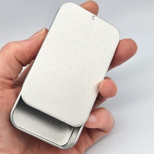 May include: A silver metal tin with a hinged lid. The tin is open and the lid is lying on top of the tin.