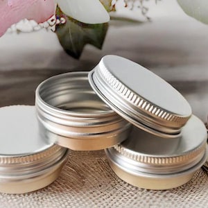 May include: Four silver metal tins with lids, stacked on a brown hessian surface. The tins are round and have a smooth, shiny finish.