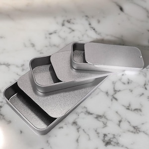 May include: Four silver metal rectangular boxes stacked on top of each other. The boxes are different sizes and are all closed.