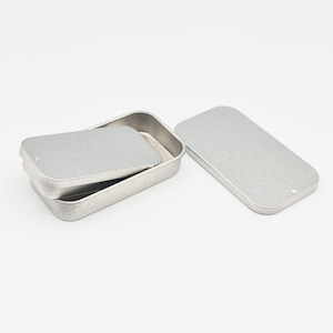 May include: A silver metal tin with a hinged lid. The tin is rectangular and has a smooth, slightly textured surface.