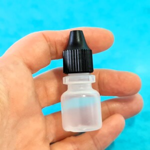 May include: A small, clear plastic bottle with a black cap and a dropper tip. The bottle is held in a hand against a bright blue background. The bottle appears to contain a clear liquid.