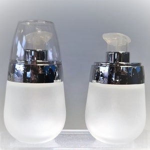 May include: Two frosted glass bottles with silver caps. The bottle on the left has a clear plastic lid. The bottle on the right has a pump dispenser.