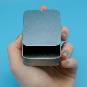May include: A silver metal tin with a hinged lid. The tin is empty and has a slightly textured surface.