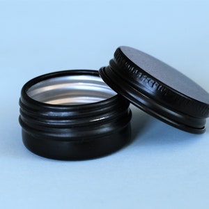 May include: A black metal tin with a silver lid. The tin is small and round, and the lid is slightly ajar.