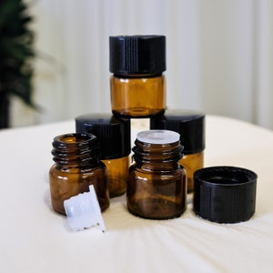 May include: A group of six small amber glass bottles with black screw-on caps. The bottles are arranged in a pyramid shape, with the top bottle being the largest. The bottles are all empty.
