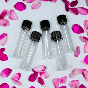 May include: Six clear glass bottles with black screw-on caps. The bottles are arranged in a semi-circle on a white surface with pink rose petals scattered around them.