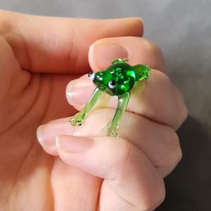 以下が含まれることがあります： 白い目をした小さな緑色のガラス製のカエルの置物。カエルは後ろ足で座り、前足を伸ばしています。