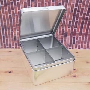 May include: A silver metal box with a hinged lid and four compartments inside. The box is open and the lid is propped up.