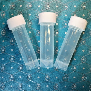 May include: Three clear plastic vials with white screw-on lids. The vials are standing upright on a blue and white patterned surface.