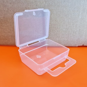 May include: A small, clear plastic hinged box, rectangular in shape, with a handle. The box is open, showing the inside. The background is orange and brown.