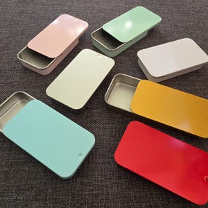 May include: A selection of rectangular metal tins in various colours, including pink, green, white, yellow, red, and teal. Some tins have their lids open, revealing the interior. The tins are arranged on a grey surface.
