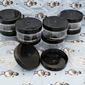May include: A set of clear plastic jars with black lids. The jars are stacked on top of each other and some are lying on their side. The jars are on a blue and white patterned fabric.