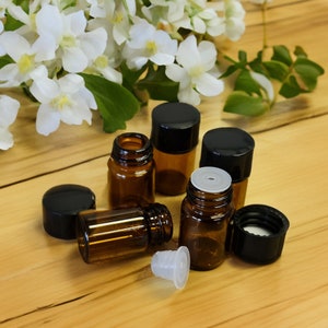May include: Six small brown glass bottles with black lids, some with white droppers, on a wooden surface. The bottles are arranged in a semi-circle with a few white flowers in the background.