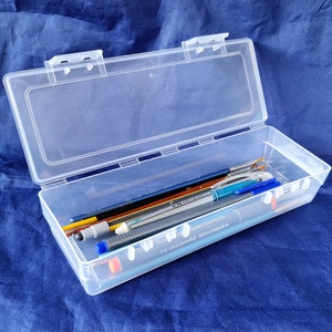 May include: A clear plastic storage box with a hinged lid, containing a variety of writing implements, including pencils, pens, and markers.