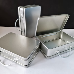May include: Three silver metal storage boxes with handles. The boxes are rectangular and have a simple design. The top box is open and the inside is visible.