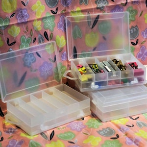 May include: Two clear plastic storage boxes with lids. The top box is open and has multiple compartments filled with colourful office supplies, such as paper clips, push pins, and staples. The bottom box has four compartments.