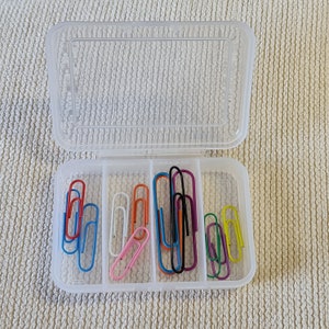 May include: A clear plastic container with multiple compartments holds a variety of colourful paper clips. The clips are arranged in rows and include blue, red, green, yellow, orange, white, and black.