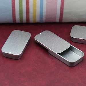 May include: Three silver metal rectangular tins with hinged lids. The tins are on a red surface.