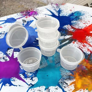May include: Five clear plastic containers with lids, some stacked on top of each other. The containers are on a white surface with colourful paint splatters.