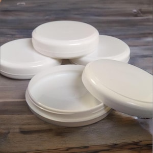 May include: Several white, round cosmetic jars with lids are displayed. One jar is open, showing the inside. The jars are made of a smooth, opaque material and are placed on a wooden surface. Suitable for storing creams, lotions, or other beauty products.
