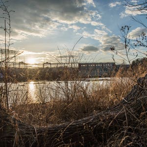 May include: A view of a large industrial building with a river in the foreground. The building is made of concrete and has many windows. The river is calm and reflects the sky. The sun is setting in the background, casting a golden glow over the scene.