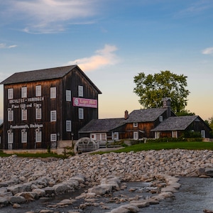 May include: A historic wooden mill building with a sign that reads "Nickless-Hininger Flour Mill and Store" and "Frankenmuth, Michigan". The building is surrounded by a rocky riverbed and a small stream.
