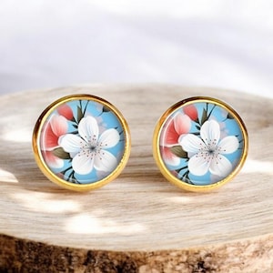 Puede incluir: Un par de pendientes de botón dorados con fondo azul y una flor blanca con detalles rosas.