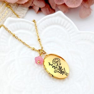 Op de afbeelding: Goudkleurige medaillonketting met bloemmotief. Het ovale medaillon heeft een zwarte bloemenillustratie en een kleine roze bedel. De ketting wordt gepresenteerd op een wit, getextureerd oppervlak, met roze bloemen op de achtergrond.