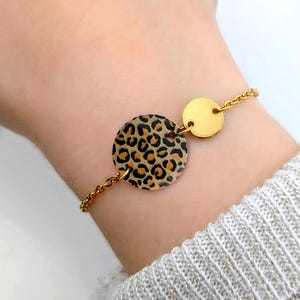 Può includere: Bracciale dorato con un grande ciondolo rotondo con stampa leopardata. Il bracciale include un disco più piccolo color oro pieno e una delicata catena. Il bracciale è mostrato su un polso con un maglione colorato.