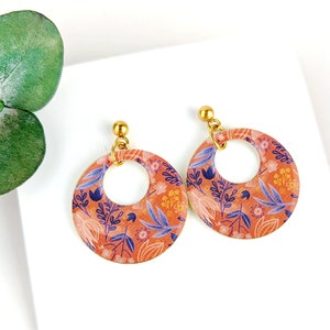 Peut inclure: Boucles d'oreilles créoles avec un motif floral orange, bleu et jaune. Les boucles d'oreilles ont des tiges dorées et une forme circulaire avec un centre évidé. Le texte "Version B" est visible en bas.