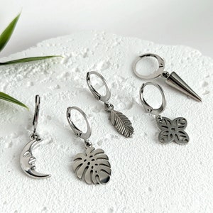 Puede incluir: Conjunto de seis pendientes de aro plateados con diferentes colgantes, incluyendo una luna creciente, una hoja, una flor, una hoja de monstera y una punta.