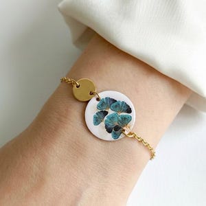 Può includere: Bracciale dorato con un pendente circolare bianco con un motivo floreale turchese. Il bracciale include un piccolo disco dorato e una catena delicata. Il gioiello è indossato al polso, mettendo in risalto il design e il contrasto cromatico.