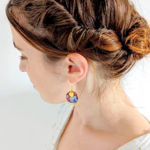 May include: Gold hoop earrings with a round, blue, red, and white patterned charm. The charm has a gold circle around it.