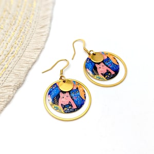 Puede incluir: Pendientes de aro dorados con un dije redondo que presenta una ilustración colorida de gatos. El dije está superpuesto a un disco dorado más pequeño.