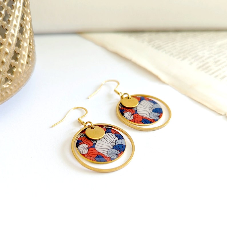 May include: Gold hoop earrings with a red, white, and blue floral pattern. The earrings have a small gold disc charm.