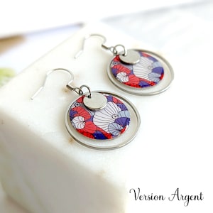 May include: Silver earrings with a round, patterned design featuring red, white, and purple colors. The earrings have a double circle design with a smaller circle in the center.