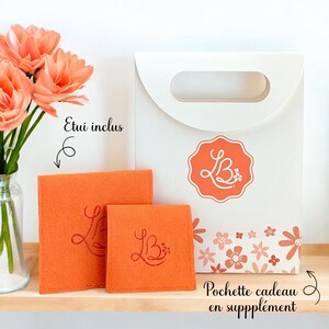 May include: A white gift bag with a floral pattern and the text "Pochette cadeau en supplement". The bag has a handle and a logo with the letters "LB" in a circle. Two orange felt pouches with the same logo are included. The text "Etui inclus" is written on the image.