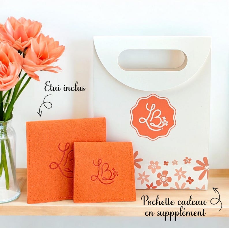May include: A white gift bag with a floral pattern and the text "Pochette cadeau en supplement". The bag has a handle and a logo with the letters "LB" in a circle. Two orange felt pouches with the same logo are included. The text "Etui inclus" is written on the image.