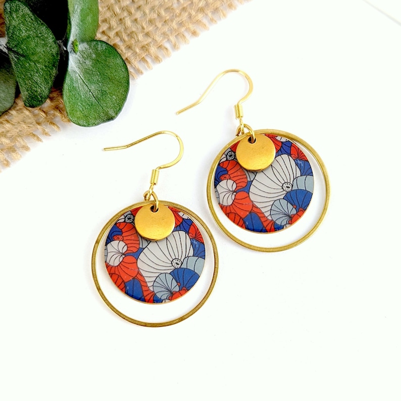May include: A pair of gold hoop earrings with a red, blue, and white floral pattern. The earrings have a smaller gold disc in the center of the floral pattern.