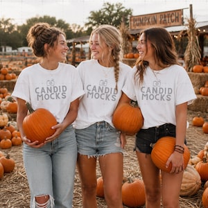 May include: Three white short-sleeve t-shirts featuring a grey camera icon and the text CANDID MOCKS printed on the front. Each shirt is worn with denim shorts or jeans, and each person holds a large orange pumpkin in a pumpkin patch setting.